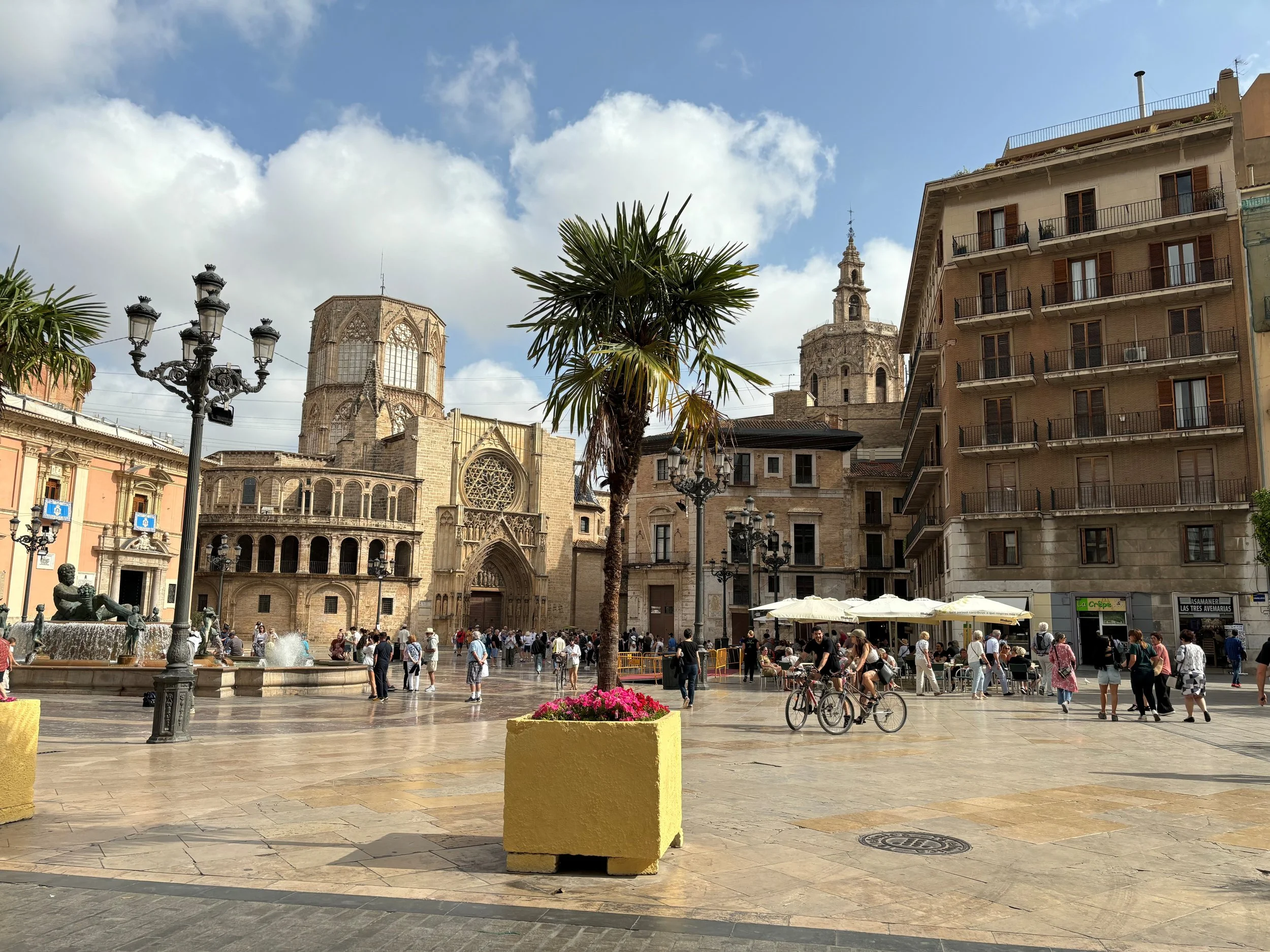 A lively city square with a large crowd of people, a stone fountain, palm trees, colorful flowers, historic churches, modern buildings with balconies, street lamps, and a clear blue sky with scattered clouds.
