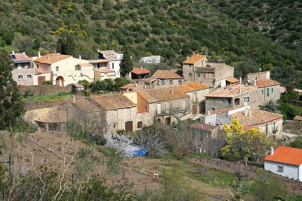 A scenic view of a hillside village with Mediterranean-style houses with red-tiled roofs, surrounded by greenery and trees.