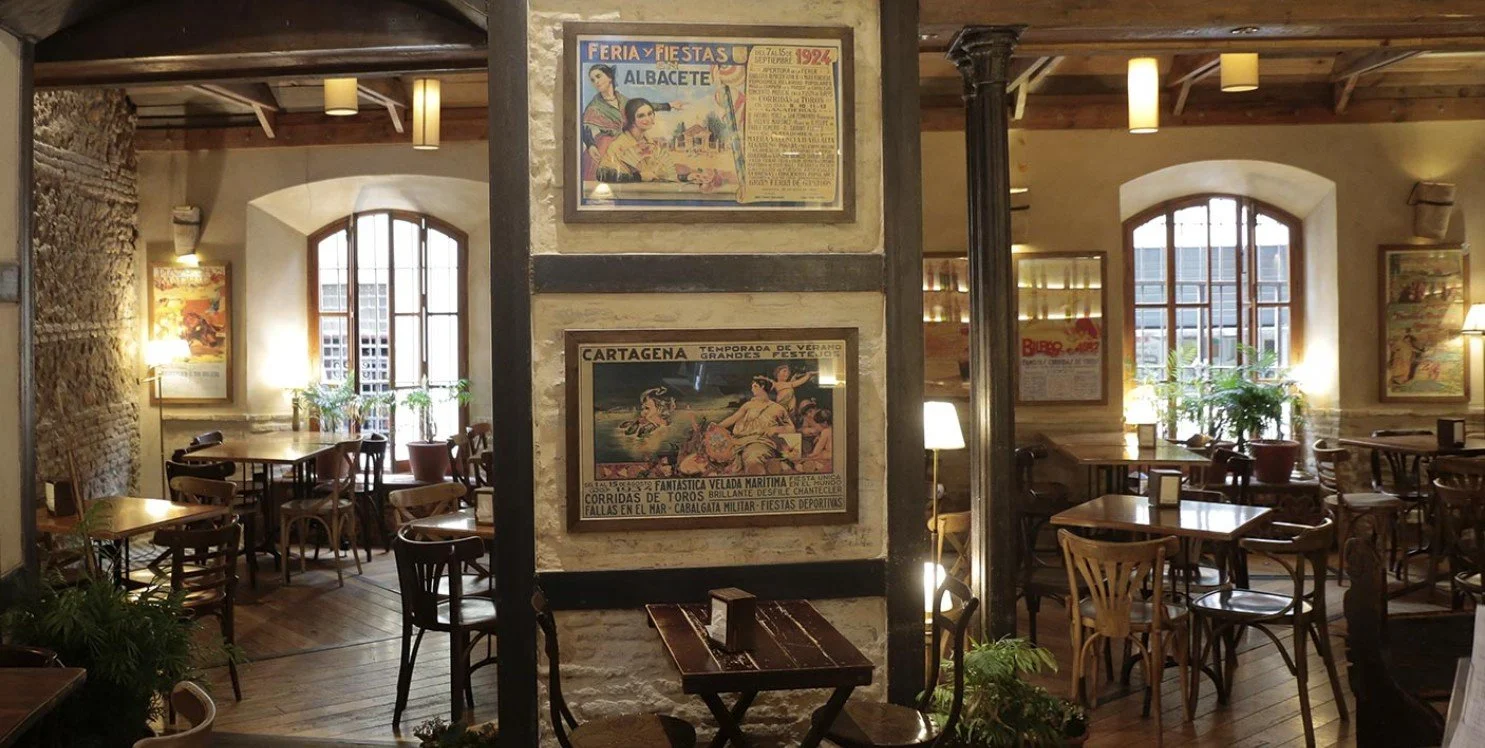 Interior of a cozy restaurant or cafe with wooden furniture, framed vintage posters on the walls, large windows, and plants, illuminated by warm lighting.