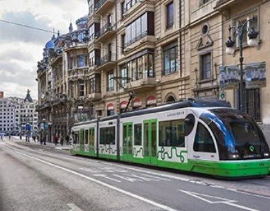 Green and white tram on city street with historic buildings and pedestrians