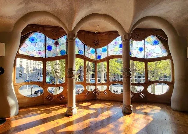 Interior view of a room with an organic, curvilinear design, large multi-pane windows with colorful stained glass, wooden flooring, and decorative columns. Outside, trees and street views are visible.