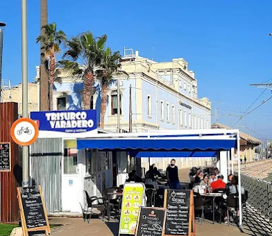 Outdoor café with people dining, yellow menu boards, and palm trees in front of a pale blue building on a sunny day.
