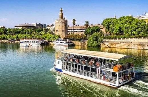 A river with a white tour boat cruising past lush green trees and historic buildings, including a tower, in a cityscape.