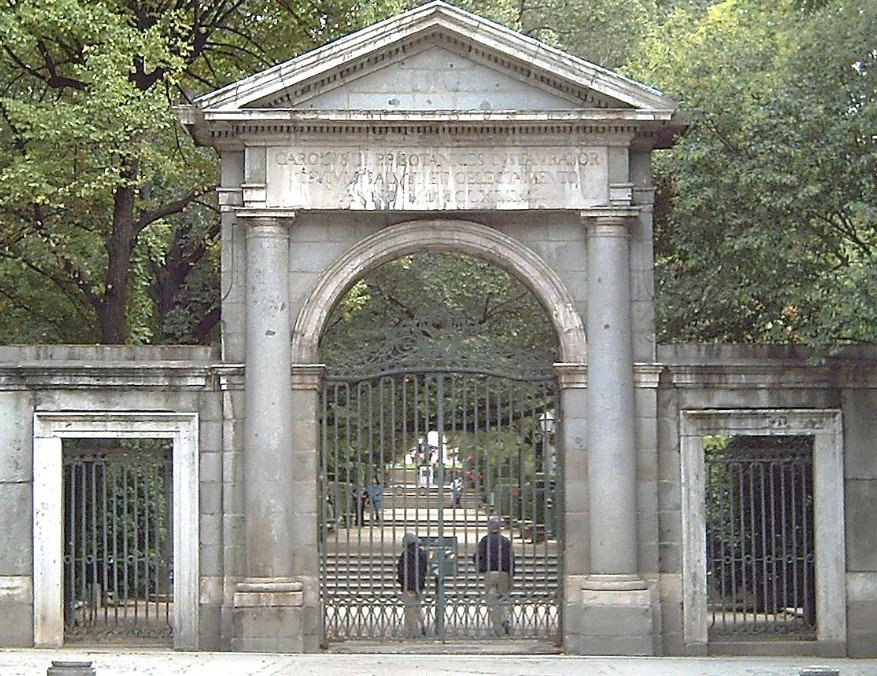 Historic stone gate with iron bars, trees in the background, and people walking inside.