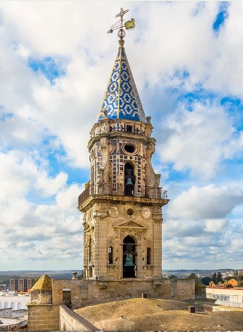 Church steeple with a blue and white tiled roof, ornate stone architecture, and a weather vane on top, against a partly cloudy sky.