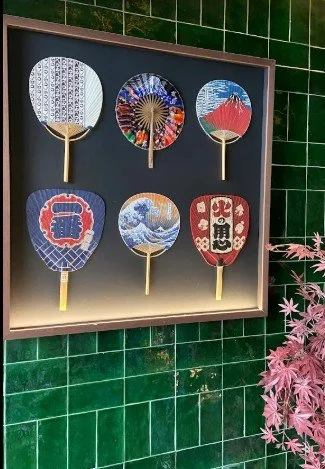 Six traditional Japanese hand fans with colorful and intricate designs displayed in a black frame against a green tiled wall, with pink foliage on the bottom right corner.