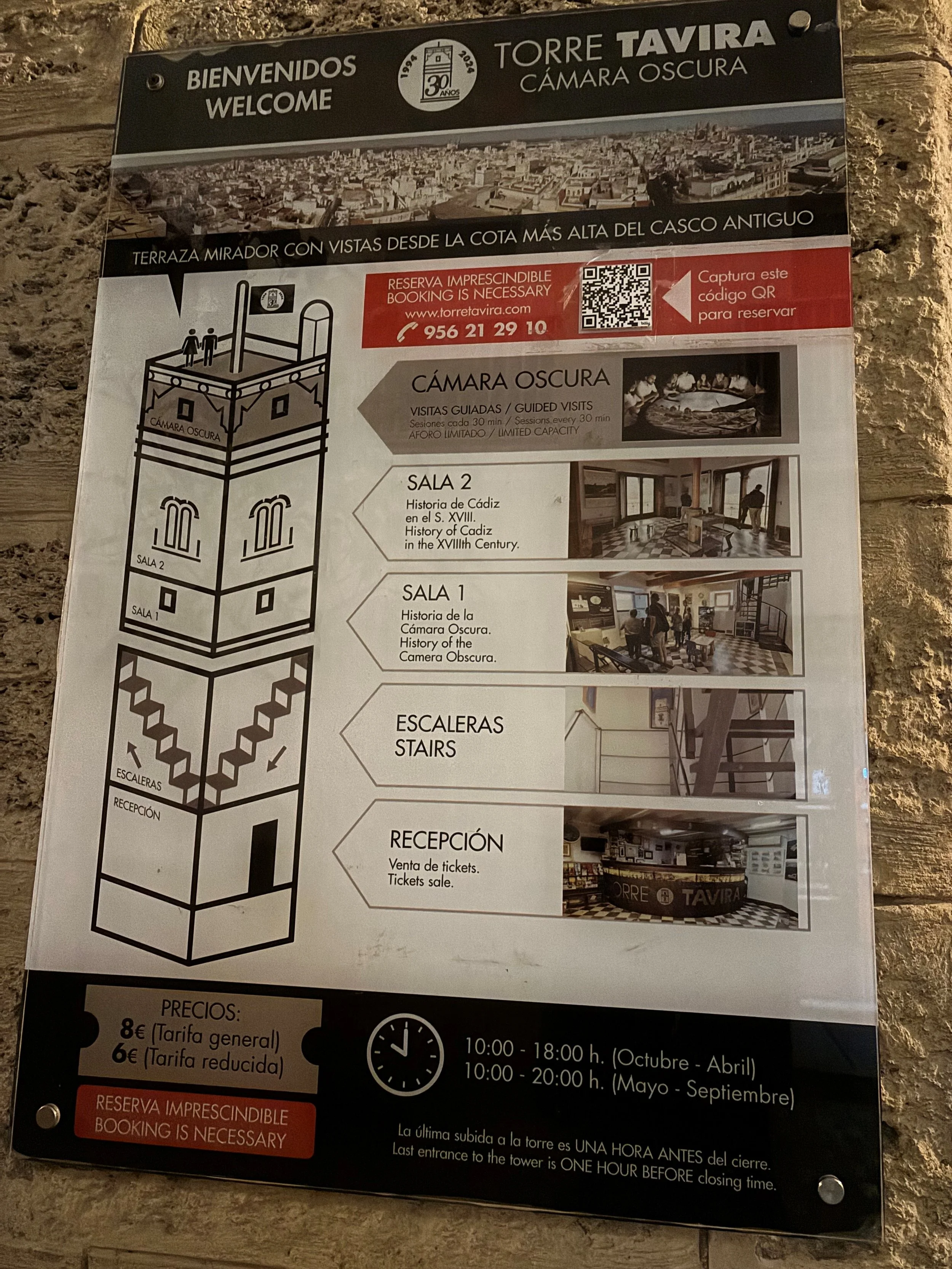 A sign describing the Torre Tavira in Cádiz, Spain. It mentions guided visits to the Camera Oscura, with photos of the interior, and information about the opening hours, ticket prices, and the importance of booking in advance.