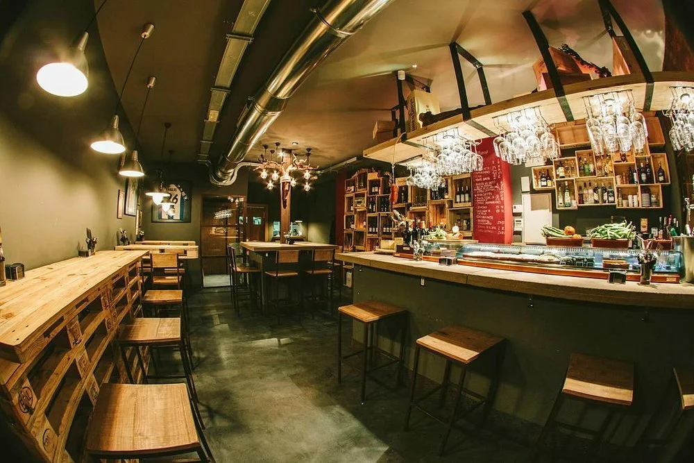 Interior of a cozy bar or restaurant with a wooden counter, shelves with bottles, hanging wine glasses, and seating areas with wooden stools and tables, dim lighting.