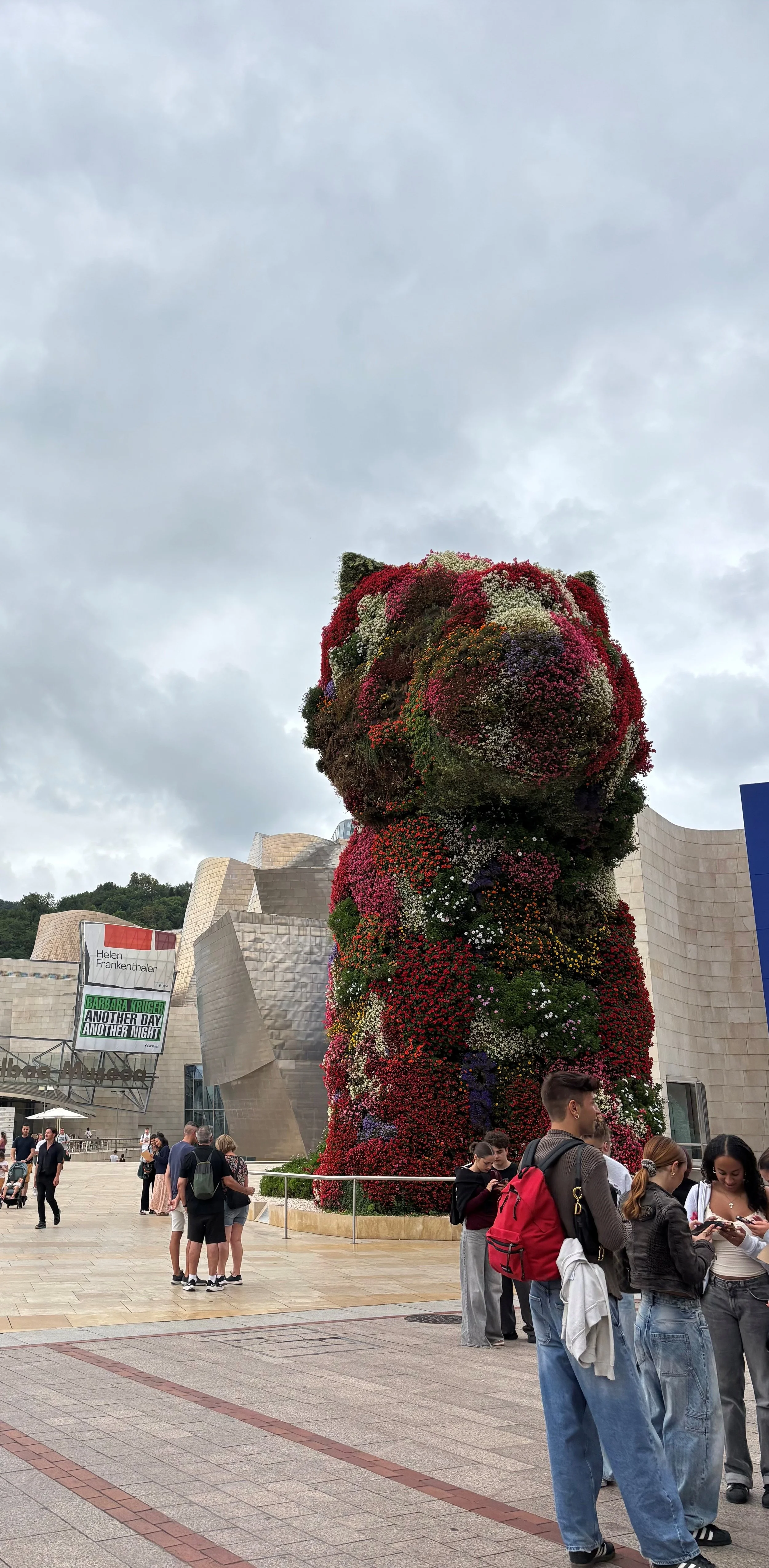 A large bear-shaped sculpture made of colorful flowers stands outside a modern building, with people gathered around observing it.