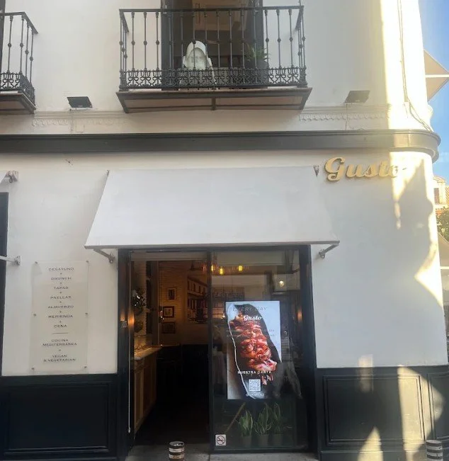 Exterior view of a restaurant with a glass door entrance, a sign listing menu items on a white wall, and the name 'Gusto' in gold letters on the upper right corner. There's a balcony with black iron railings above the entrance.
