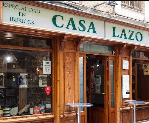 Exterior of a restaurant named Casa Lazo with a sign indicating Spanish cuisine and Iberian specialties, featuring wooden framing and two small outdoor tables.