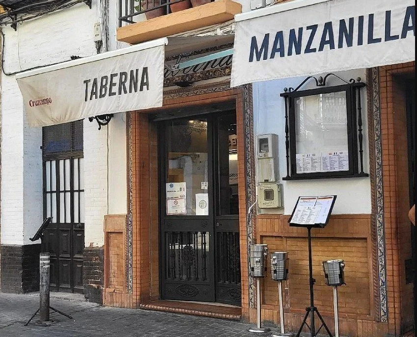 The exterior of a restaurant with two white awnings labeled 'Taberna' and 'Manzanilla', a black door, a menu stand, and some utility boxes mounted on the wall.