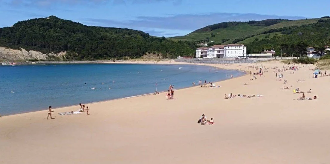 People relaxing and walking on a sandy beach with calm water, surrounded by green hills and a few buildings under a blue sky.