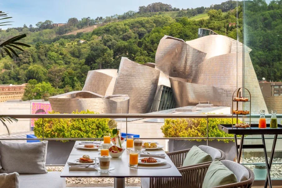 Breakfast table with orange juice, coffee, and pastries, with a view of the Walt Disney Concert Hall and green hillside through a large window.