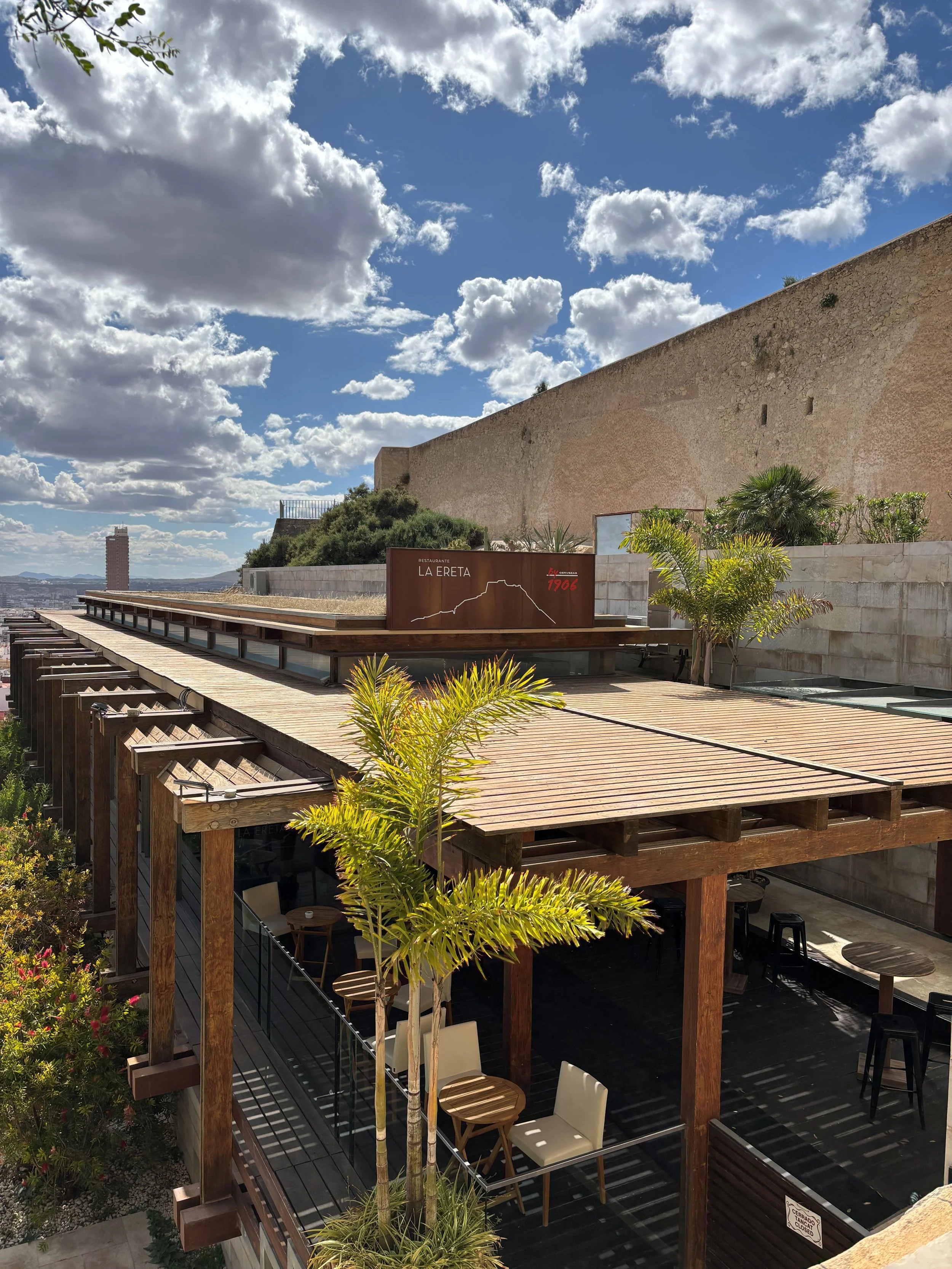 Outdoor seating area of a restaurant called La Erreta with wooden terraces, greenery, and a large stone wall under a partly cloudy sky.