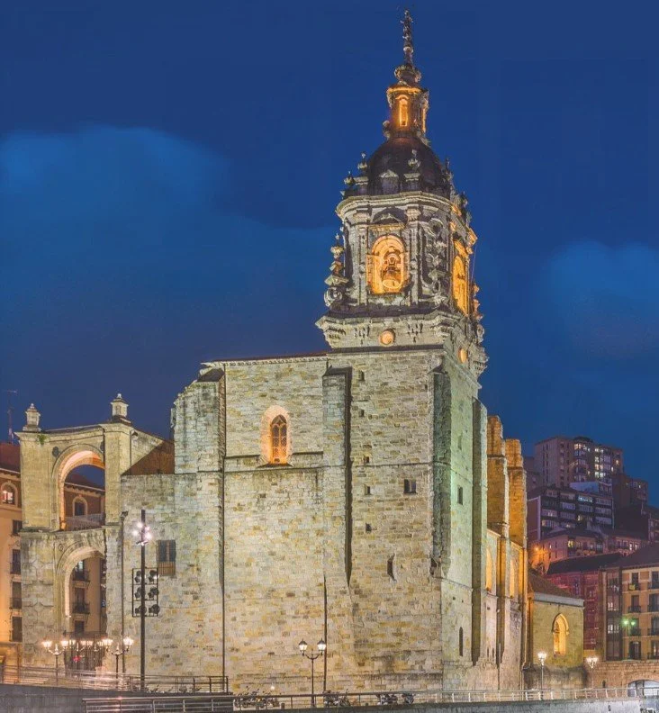 A historic church with a tall illuminated bell tower at night, with city buildings in the background.
