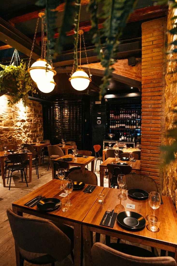 A cozy restaurant interior with wooden tables set with black plates, wine glasses, and black cutlery, exposed brick walls, hanging lamps, and lush greenery.
