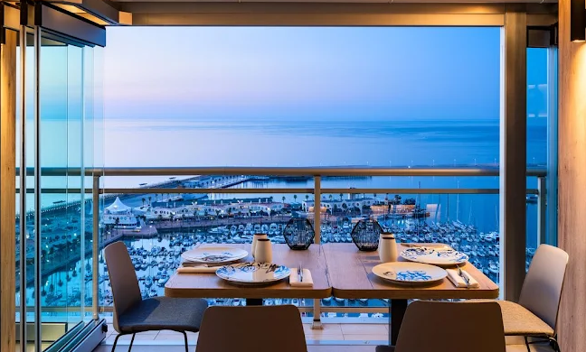 Dining table set for four with dishes, cups, and decorative lanterns, overlooking a marina and beach from a high-rise balcony at sunset.