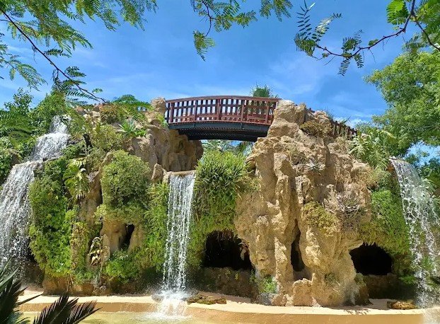 Artificial rock formation with waterfalls, a wooden bridge, lush green plants, and a blue sky.