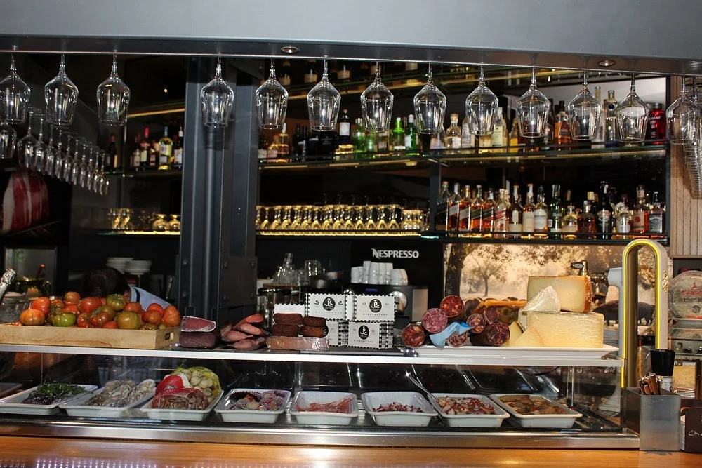 Bar counter with tomatoes, meats, cheeses, and salads, with liquor bottles, glasses, and a coffee machine in the background.