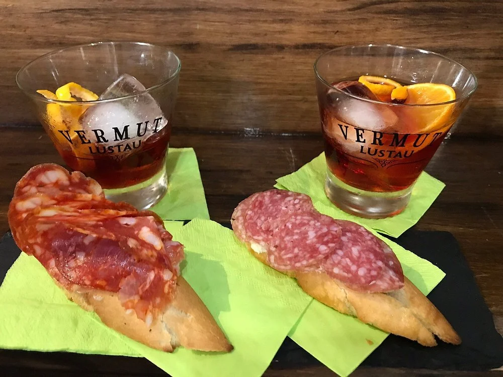 Two glasses of dark red vermouth with lemon twists and ice, placed on a wooden table. Two slices of baguette topped with cured meats, likely salami and prosciutto, on yellow napkins.