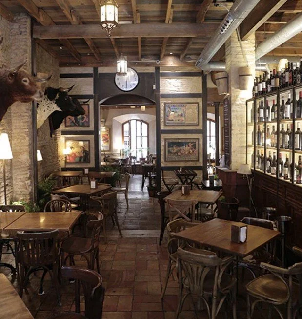 Interior of a rustic restaurant with wooden tables and chairs, a large clock on the wall, mounted animal heads, and framed artwork, with a view into an adjoining room and bottles displayed on a glass shelving unit.