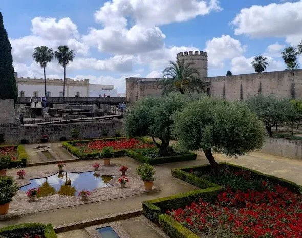 Historic fortress wall with a tower, overlooking a garden with trees, a pond, and flower beds, under a partly cloudy sky.