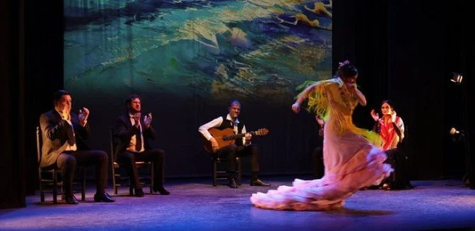A stage performance with four seated men, one playing guitar, and two women dancing, set against a colorful abstract background.