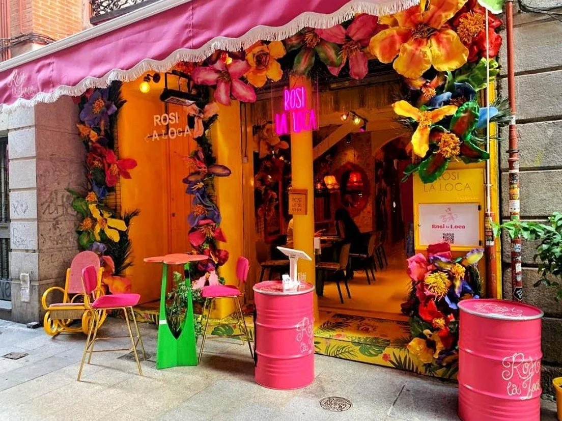 Decorated storefront with pink, yellow, and floral accents for a restaurant or cafe called 'Rosi La Loca'. Outdoor seating with pink chairs, a small green and pink table, and pink barrel tables. Colorful flowers and plants surround the entrance, with a neon sign and interior lights visible inside.