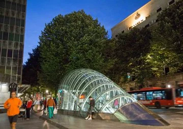 People walking past a glass underground subway entrance with a tunnel design in an urban area at dusk, trees and buildings in the background.