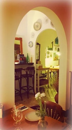 Indoor dining area with a table set with a vase of white flowers, glassware, and plates, and an arched doorway leading to another room with wall art and furniture.