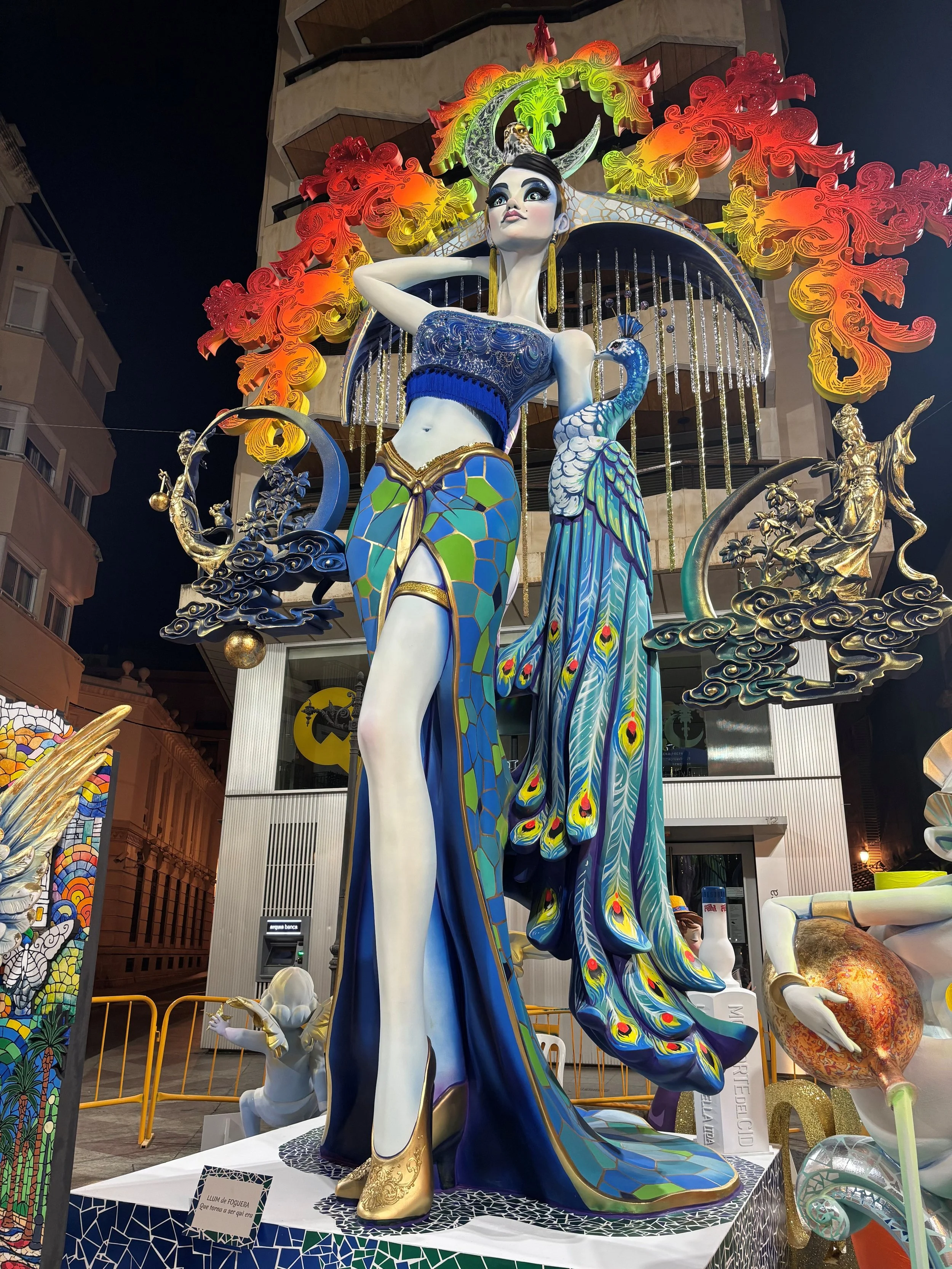 A colorful sculpture of a woman with long black hair, dressed in a blue and green outfit with peacock feather details, a peacock beside her, and decorative flames or cloud-like designs in red, orange, and yellow behind her. The sculpture is displayed outdoors at night with buildings and security barriers in the background.