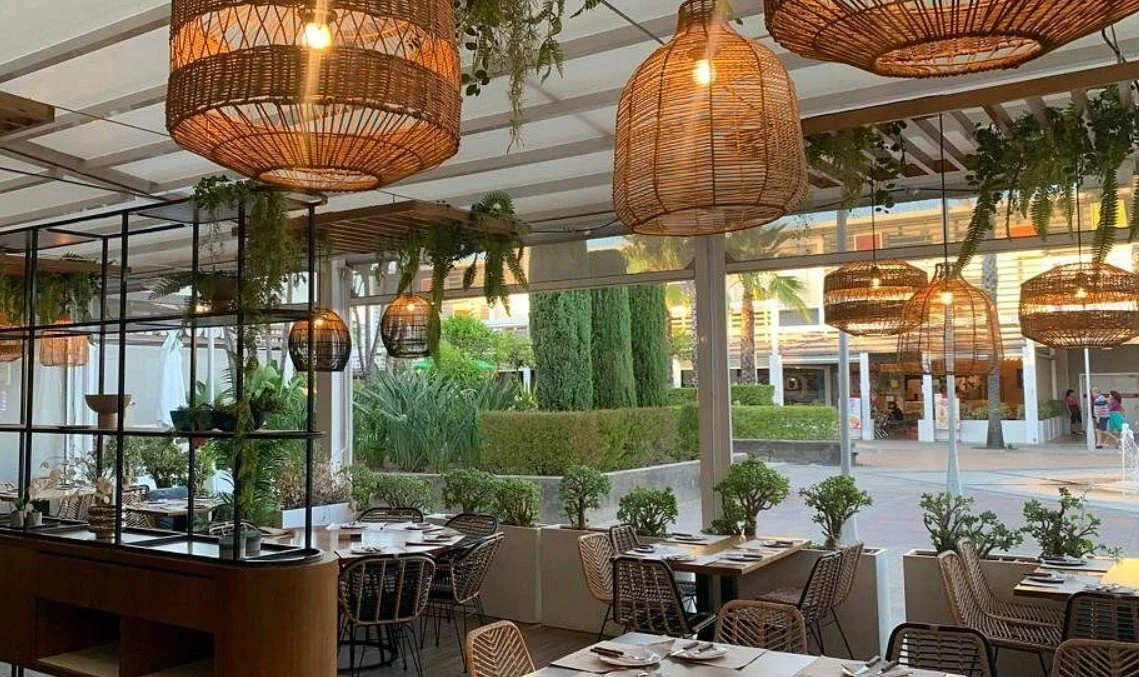 Interior of a restaurant with wicker pendant lights, wooden tables, and rattan chairs, decorated with greenery and large windows showing an outdoor street scene.