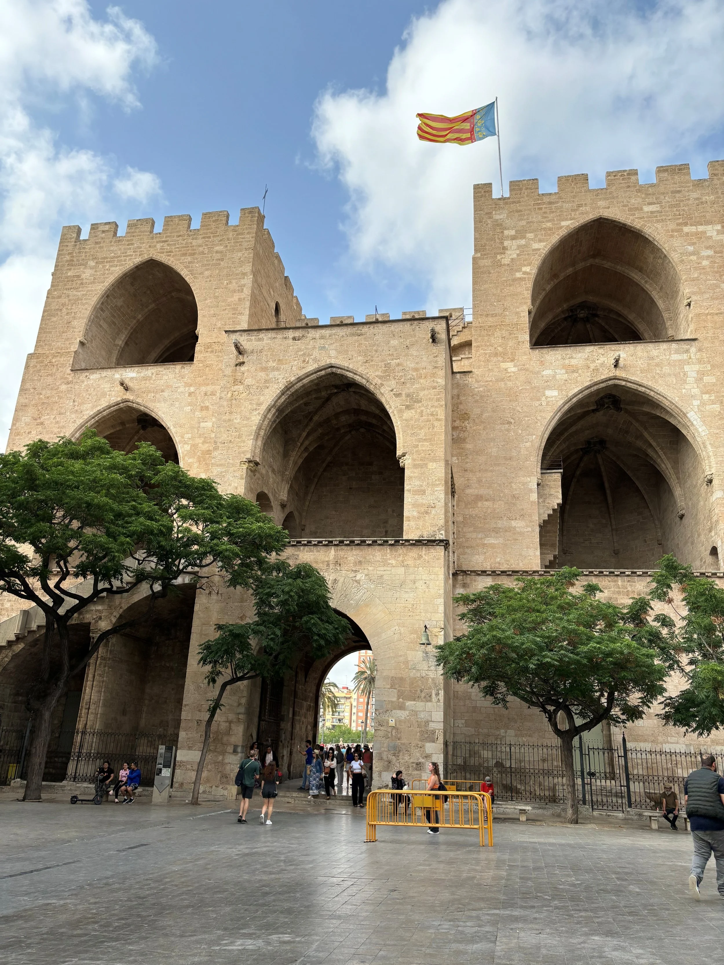People walking and sitting under a historic stone gate with two large arched openings, flanked by trees, with people visible through the arches and in the background.