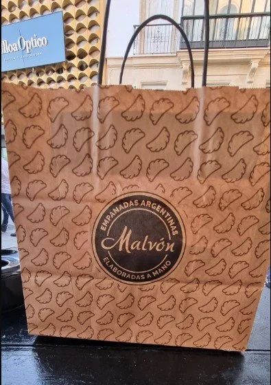 A brown paper shopping bag with a repeating pattern of hand icons and a circular logo that reads "Empanadas Argentinas Malvón" on the front.