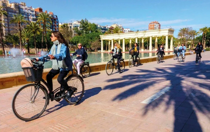 Group of people riding bicycles along a waterfront promenade with a fountain, palm trees, and a white pavilion in the background on a sunny day.