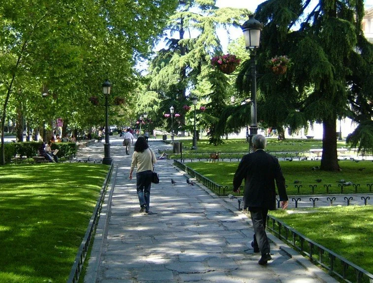 People walking along a park path lined with trees and benches, with lampposts and hanging flower baskets.