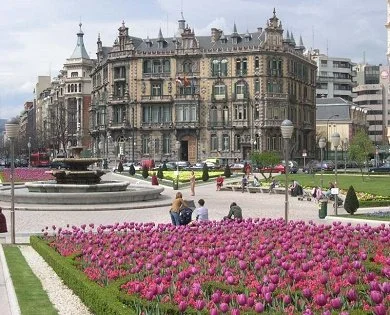 A historic building with ornate architecture in a city square, surrounded by pink tulip flowers and people sitting on benches near a fountain.