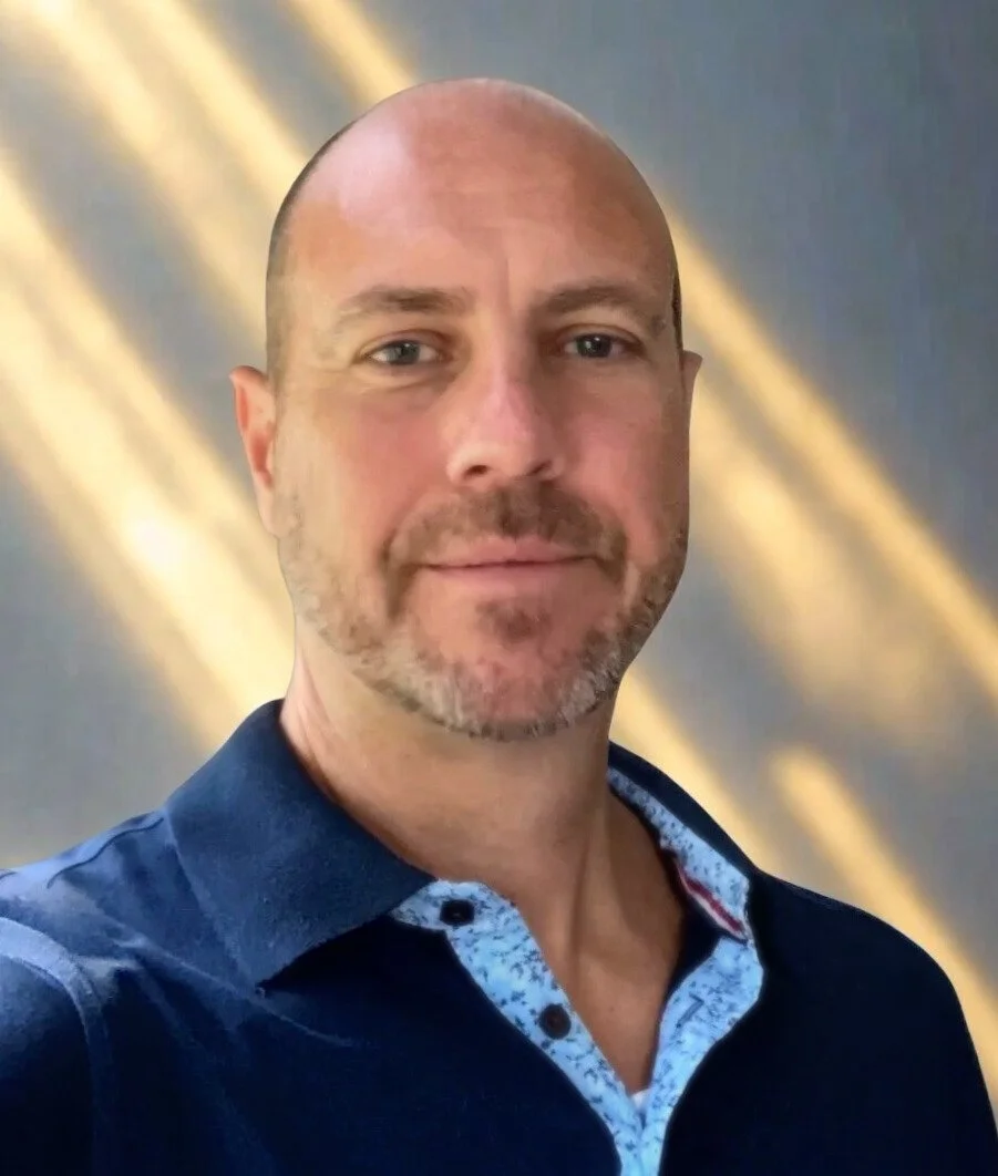 A man with a shaved head and facial hair, wearing a dark blue collared shirt with a patterned inner lining, standing in front of a gold and navy background.
