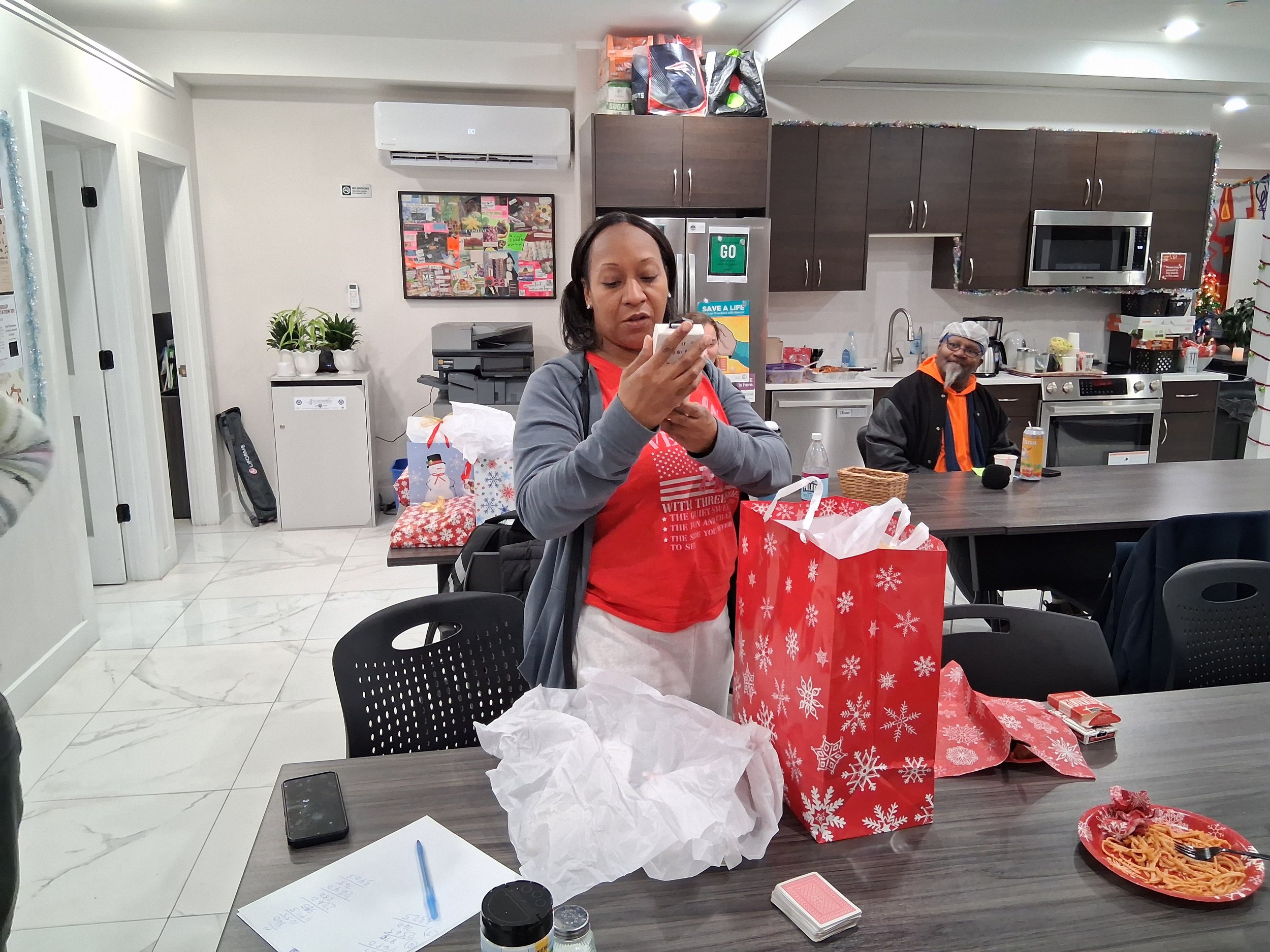 A woman in a red shirt and gray hoodie standing at a table with Christmas gift bags, tissue paper, a plate of spaghetti, and a deck of cards, with a man in a black jacket with orange hoodie in the background in a kitchen decorated for Christmas.