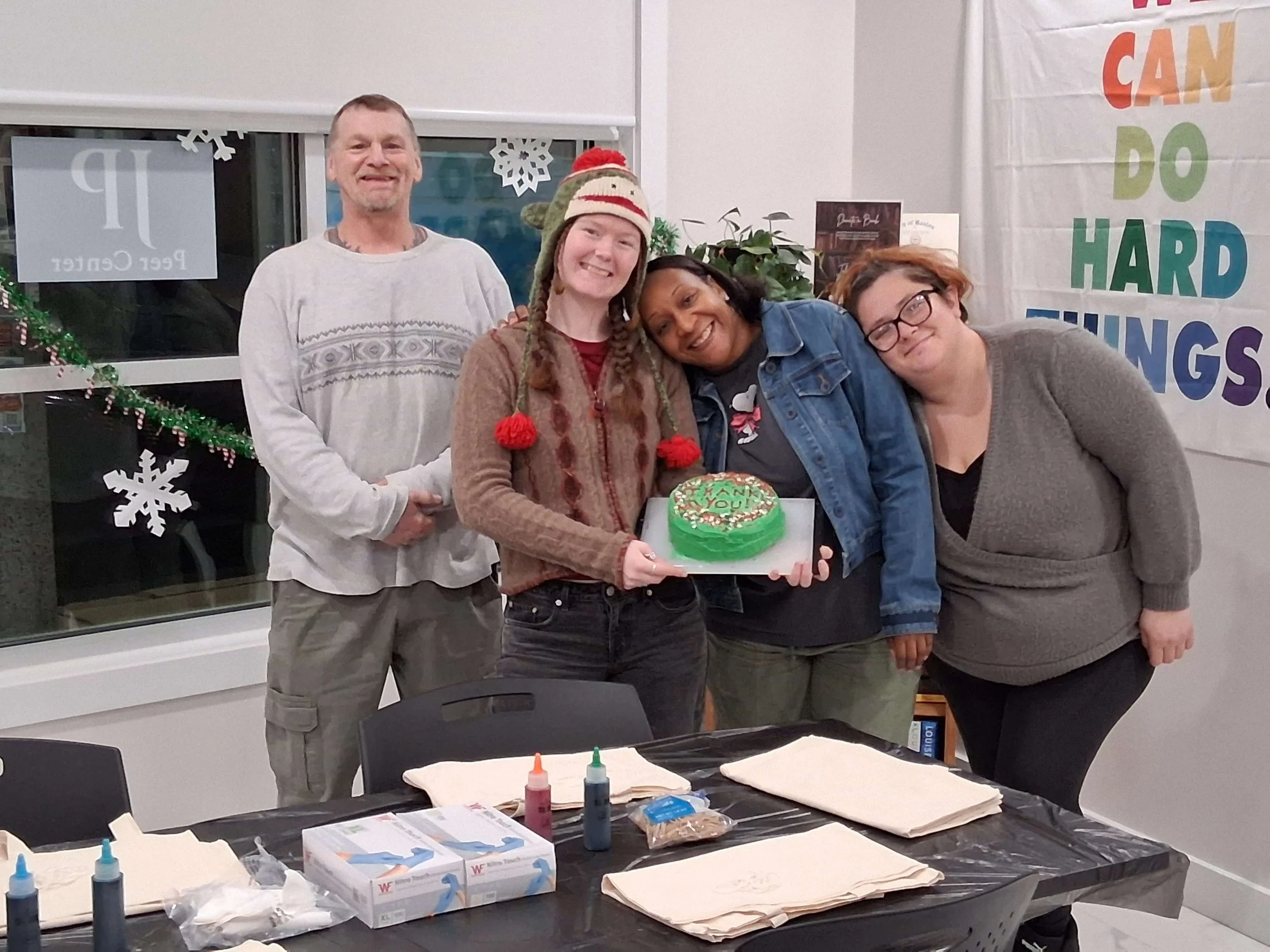 Group of five people celebrating, with a woman holding a decorated birthday cake. They are smiling and standing indoors, with Christmas decorations and a colorful sign in the background.