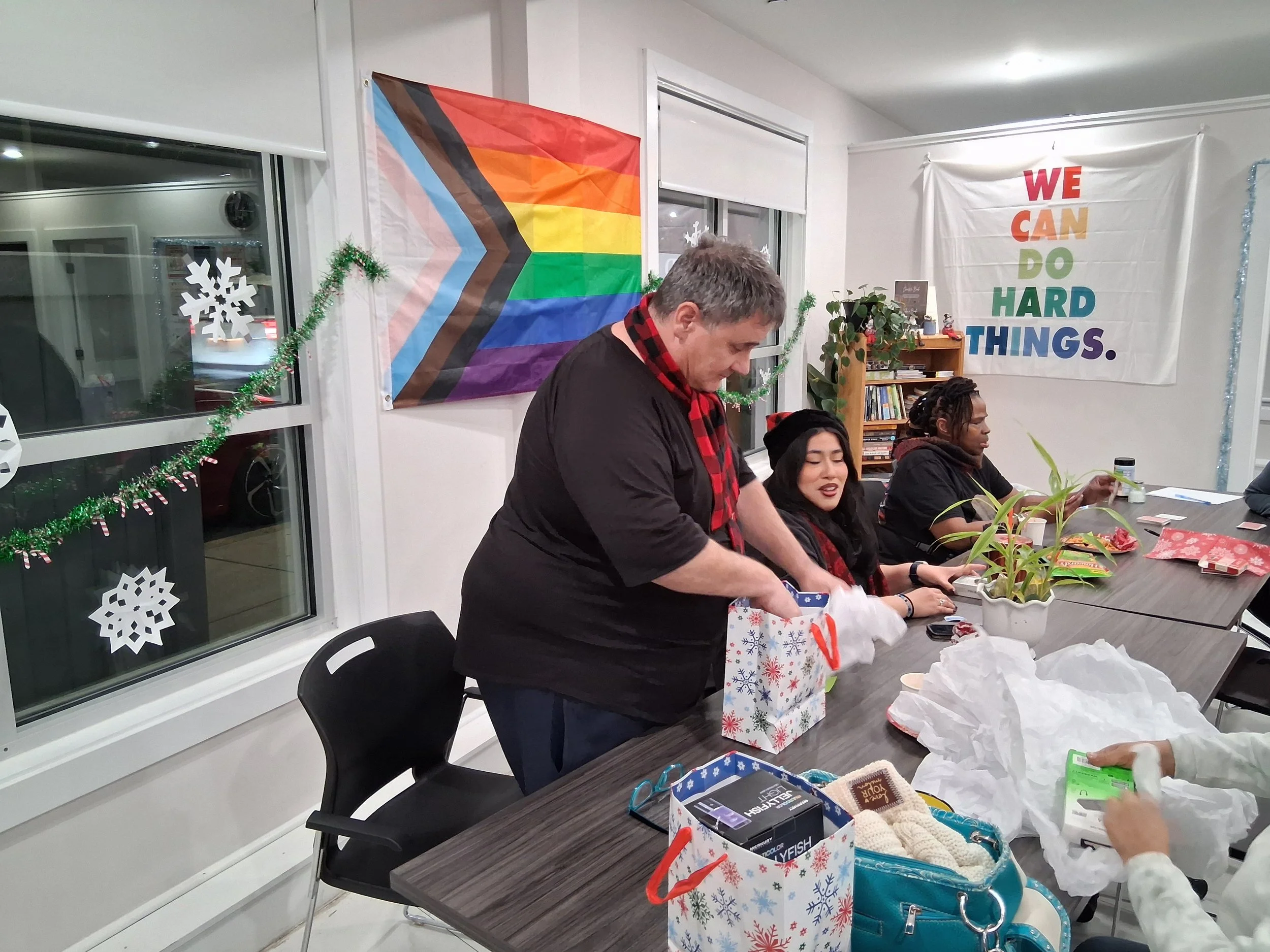 People gather around a table exchanging gifts at a holiday celebration. Decorations include paper snowflakes, a rainbow flag, and a banner that reads 'WE CAN DO HARD THINGS.'