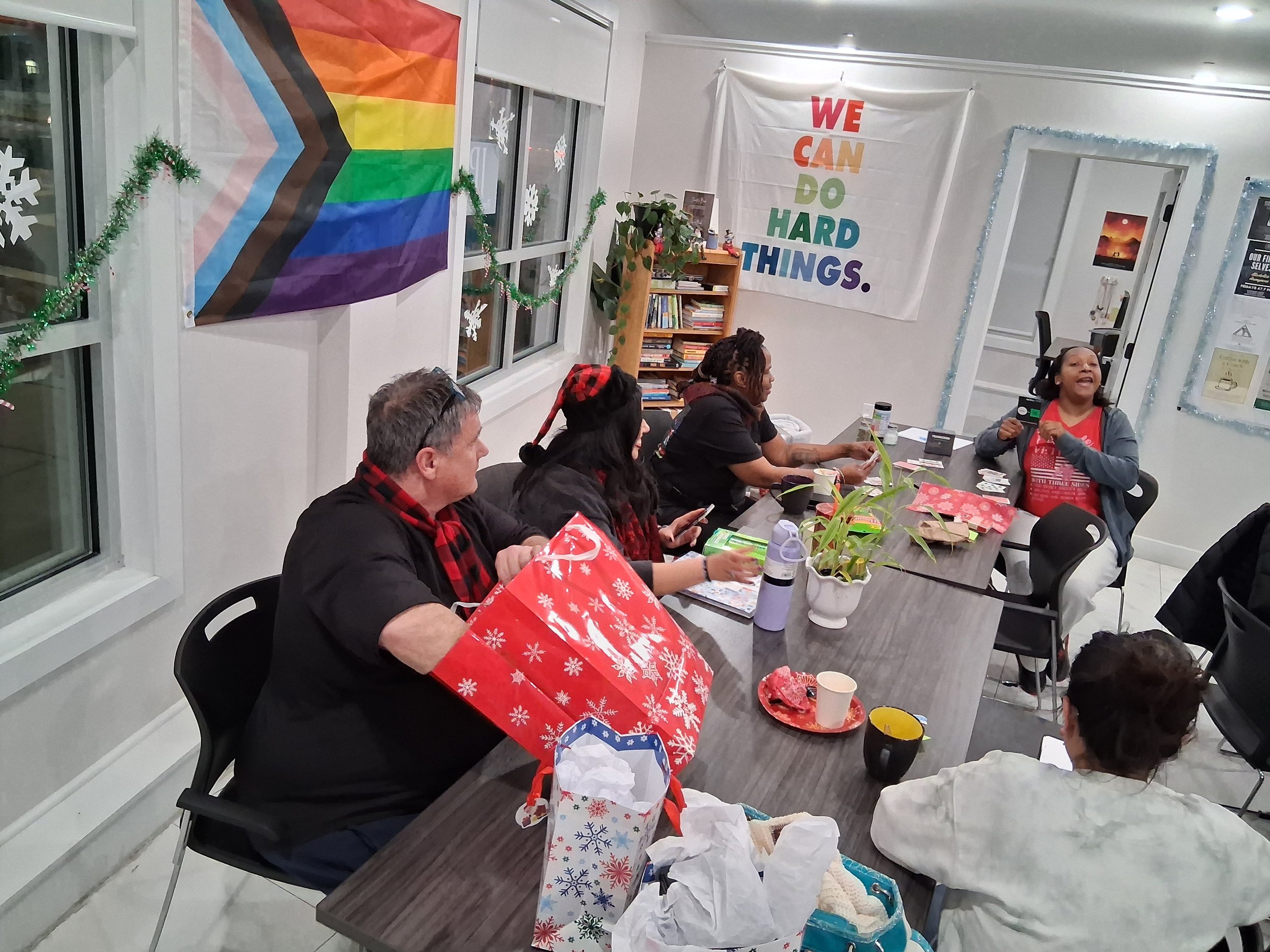 People sitting around a conference table decorated for the holidays, exchanging gifts. The room has festive decorations, a rainbow flag, and a sign that says 'We Can Do Hard Things.'
