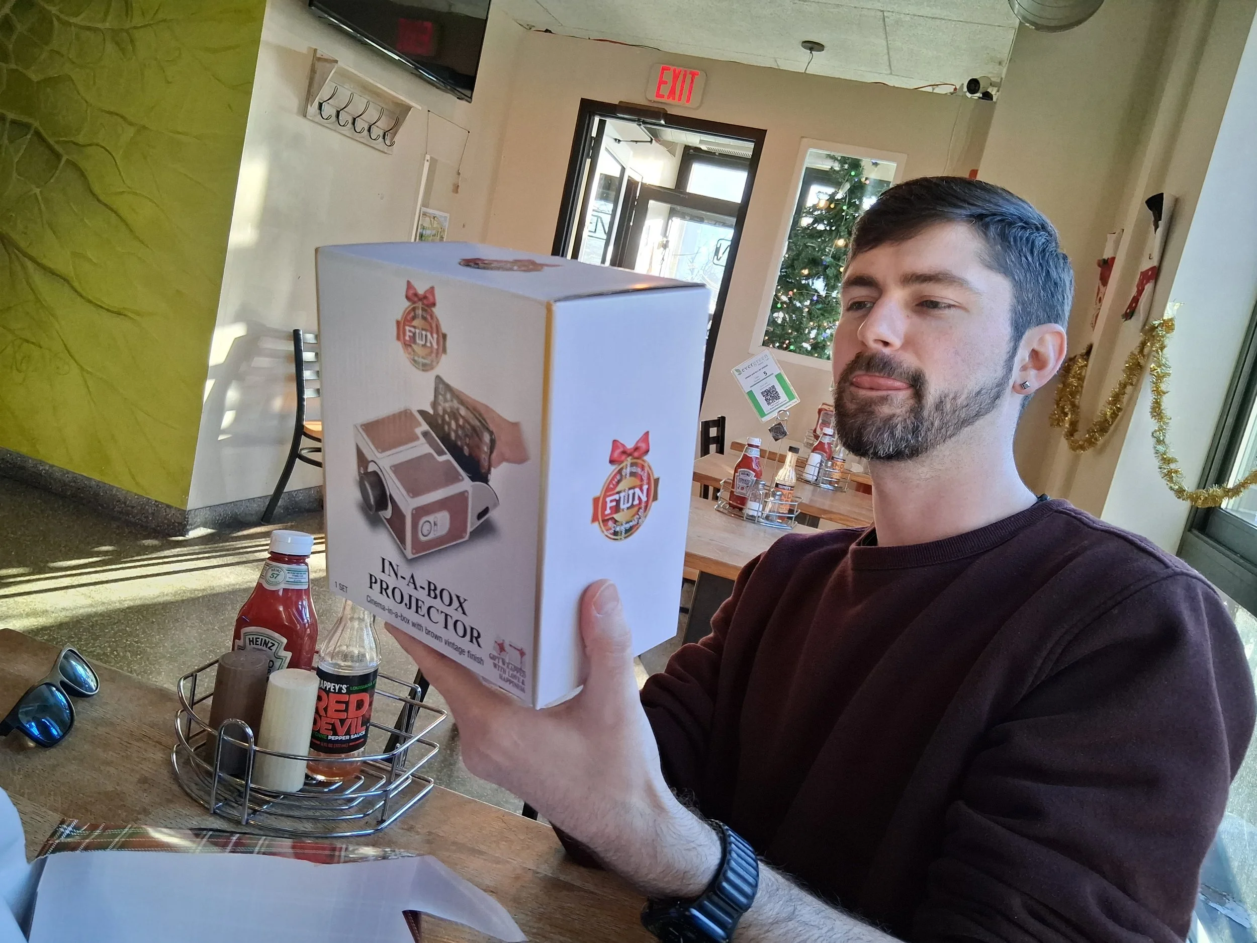 A man with dark hair and a beard is holding a box labeled 'In-A-Box Projector' in a restaurant or cafe with festive decorations, including a decorated Christmas tree in the background.
