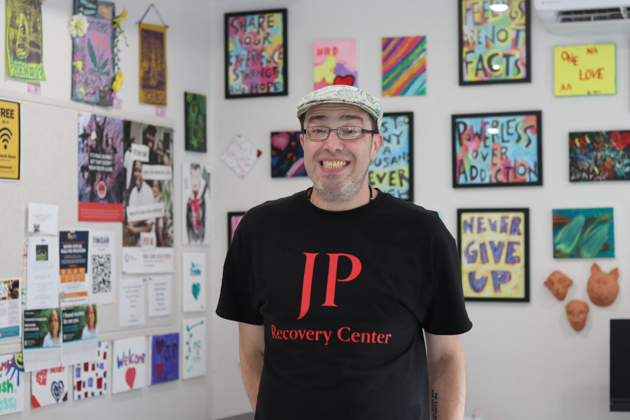 A man smiling with glasses and a cap standing in front of a wall decorated with colorful artwork, posters, and motivational quotes at a recovery center.
