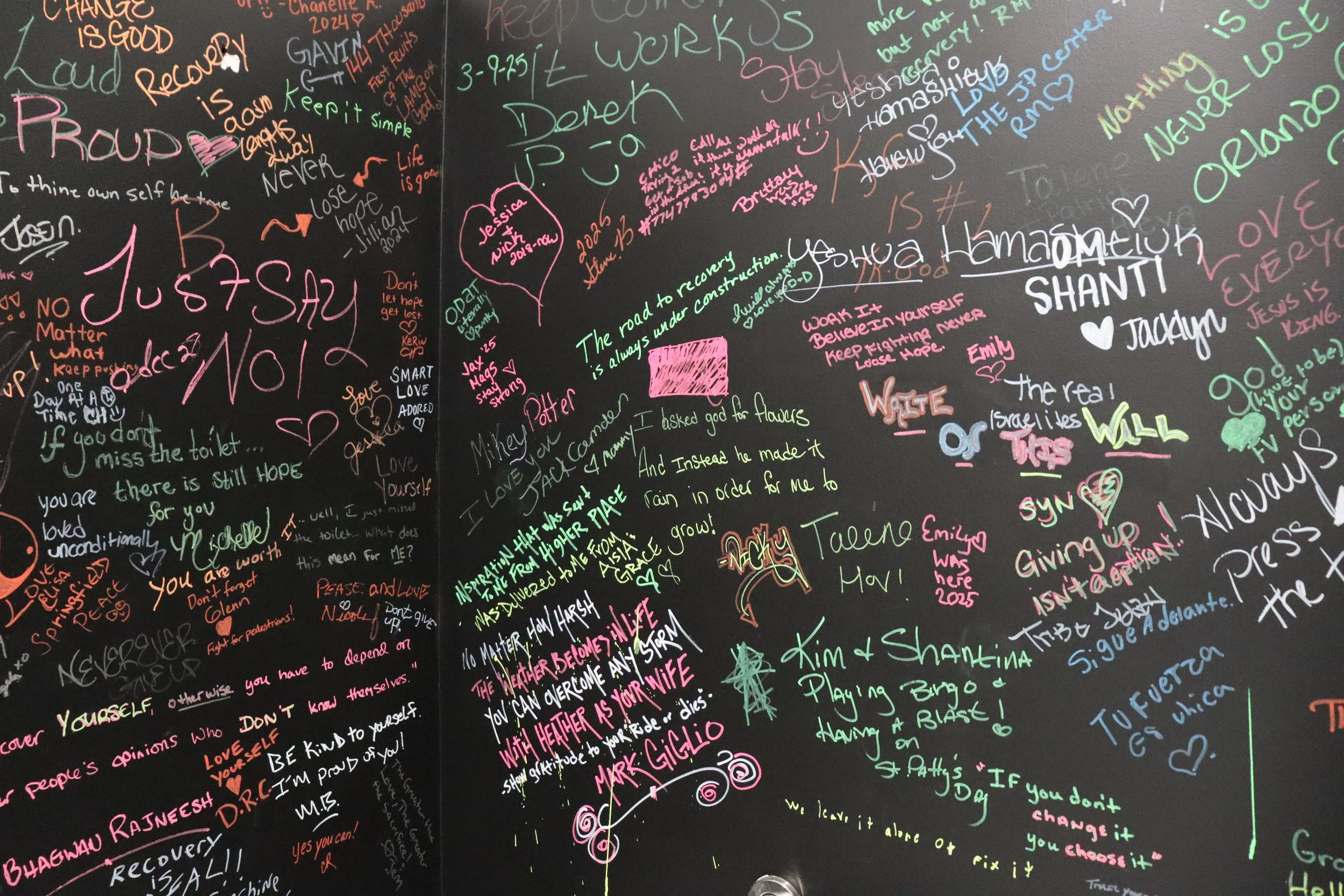 Colorful handwritten messages and quotes on a black wall, including names, motivational phrases, drawings, and signatures in pink, green, blue, yellow, and orange.