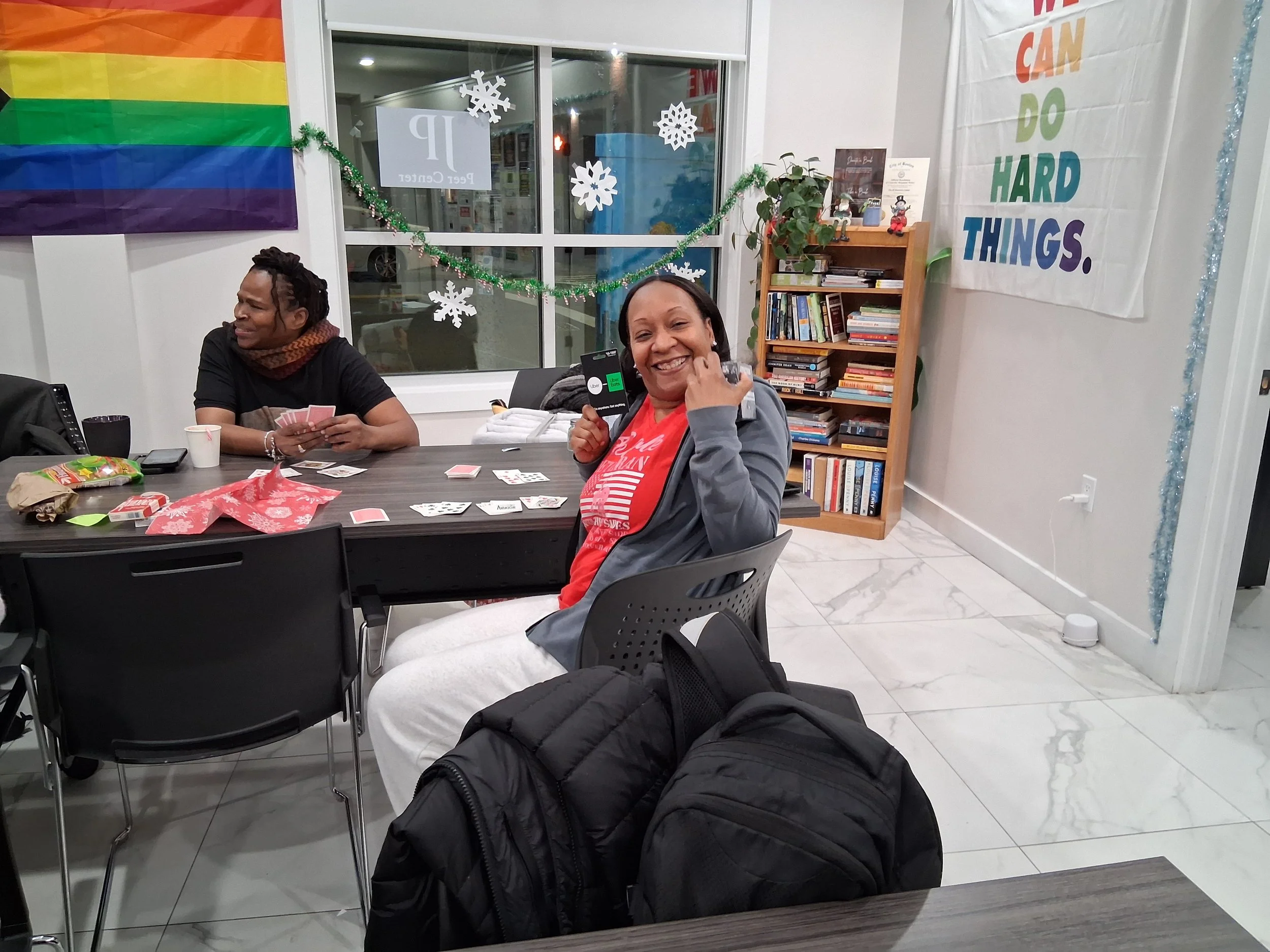 Two women are sitting at a table, playing cards and smiling. One woman, in a red shirt and gray jacket, is holding a card and smiling at the camera. The other woman, in black and a scarf, is also holding cards and looking to her left. The table has s