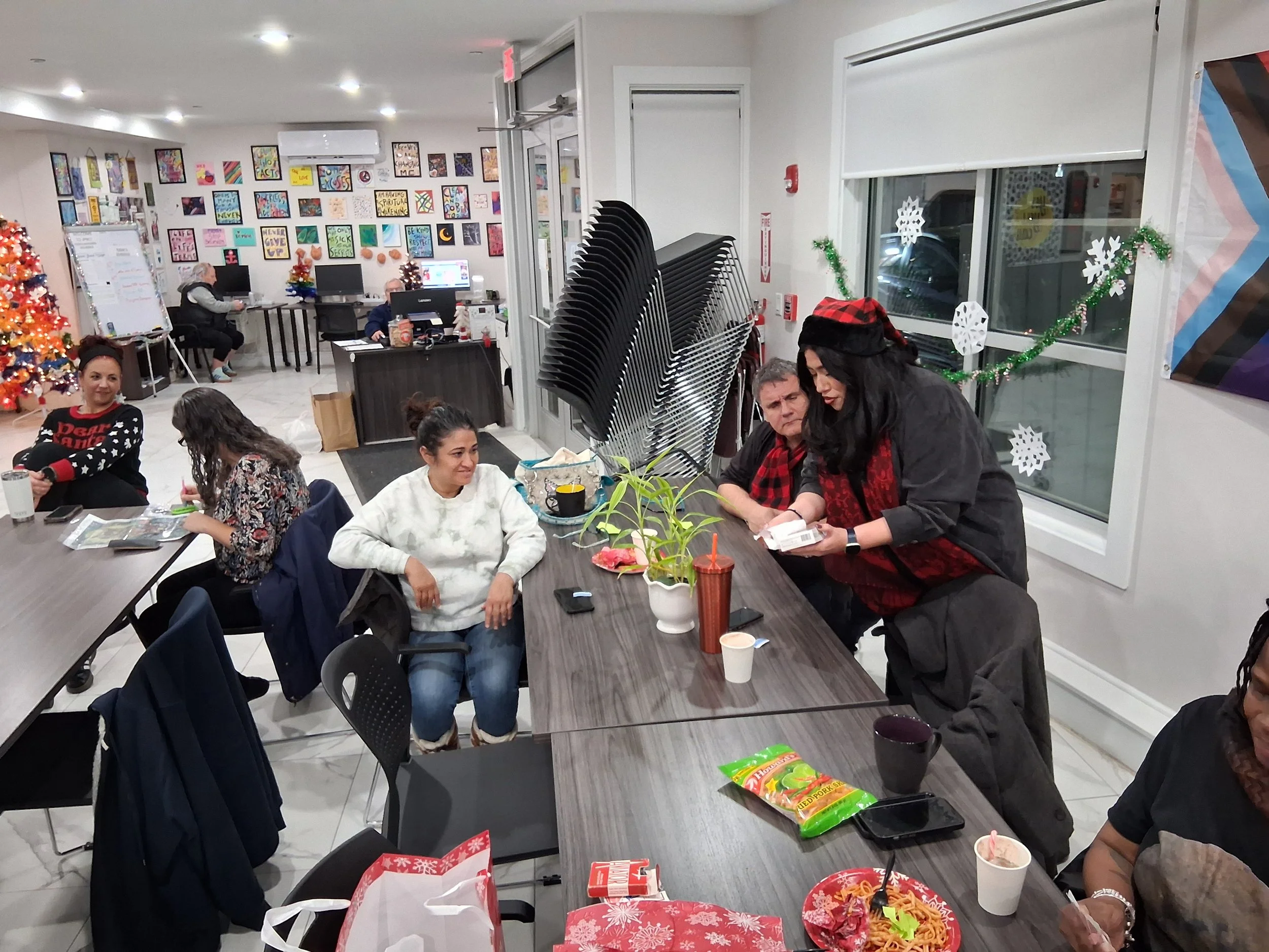People gathering in an office decorated with Christmas decorations, including a Christmas tree, snowflakes on the window, and tinsel. Some are sitting and eating, while others are standing and talking.