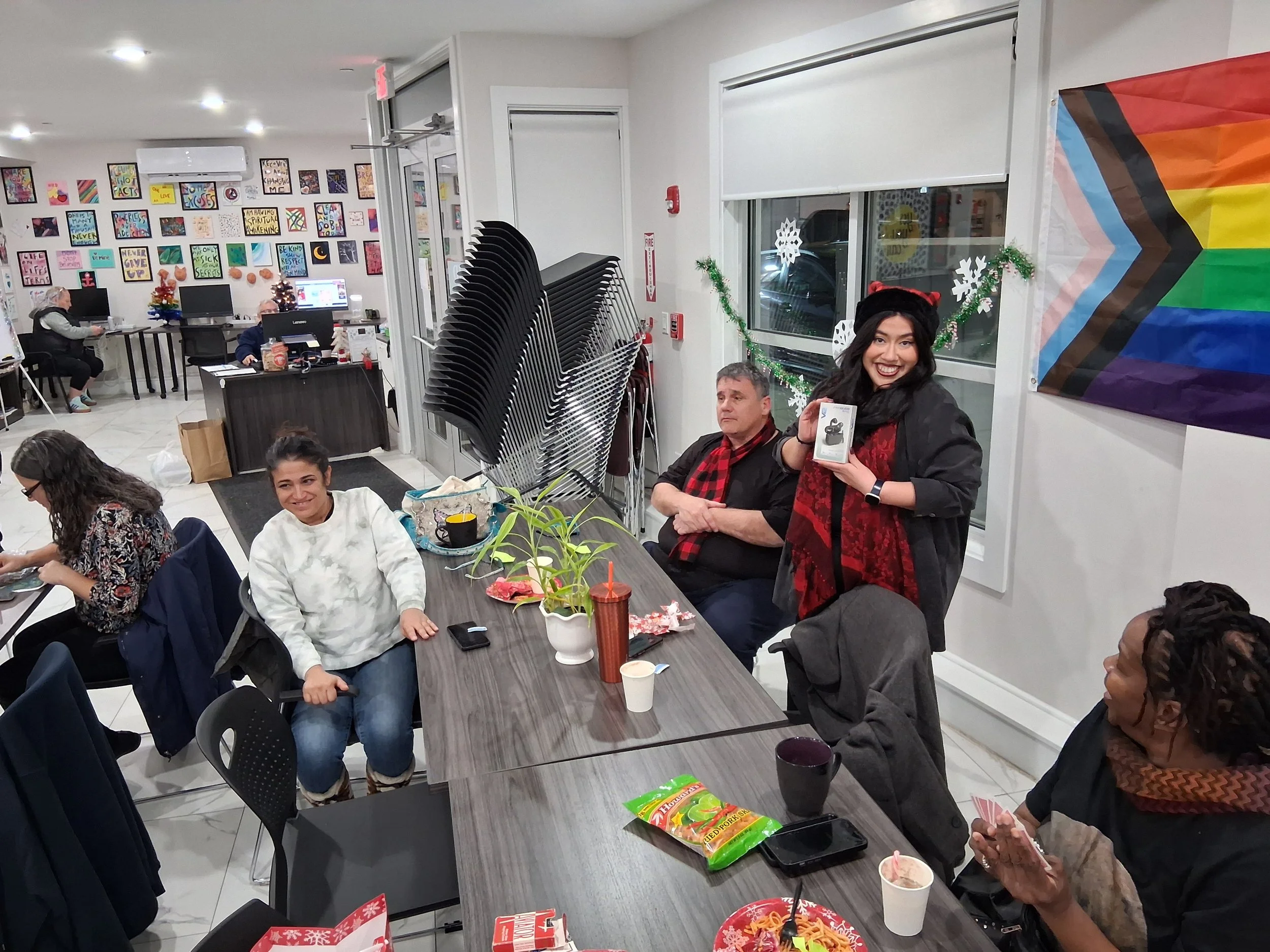 A group of diverse people gathered in a decorated room, celebrating a holiday or special event. Some individuals are sitting at a table with snacks and drinks, while a woman standing is smiling and showing a gift. The room has holiday decorations, in