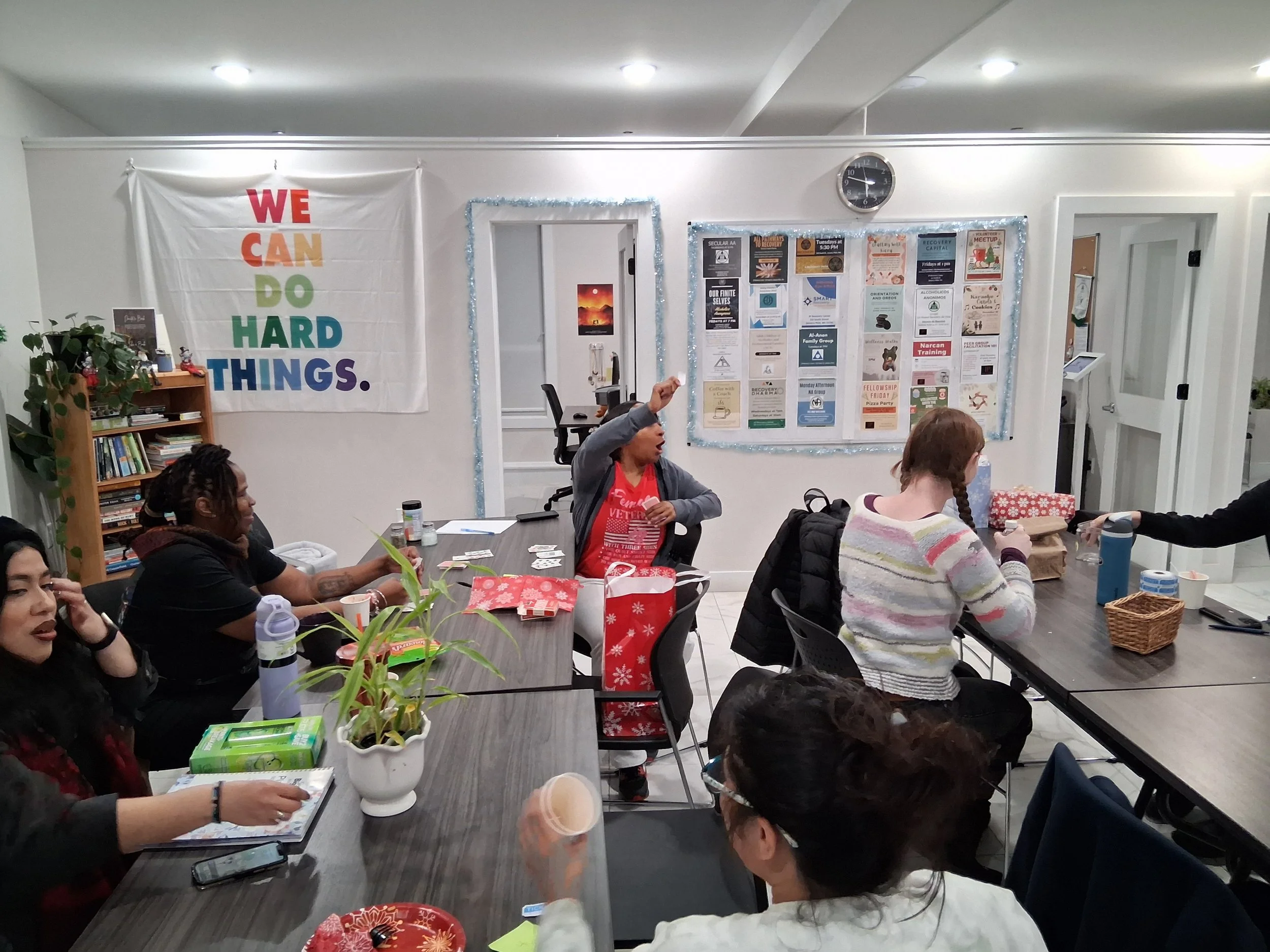 People sitting around a table in a room decorated for the holidays, with gift bags and snacks, one person in the middle raising her hand, a large rainbow-colored sign on the wall reading 'WE CAN DO HARD THINGS,' and a bulletin board with flyers.